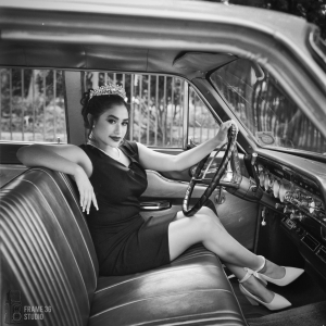 Quincea&ntilde;era in a formal dress sitting in the driver's seat of a car, smiling at the camera