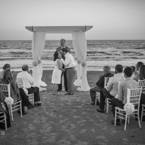 Bride and groom kissing beneath a wedding arch on the beach as guests applaud